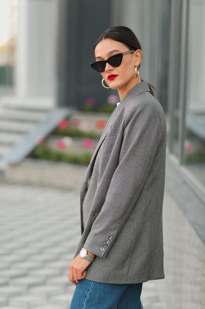 Attractive stylish woman in sunglasses wearing grey jacket posing with red lips outdoor in city Goodlooking stylish lady walking in city