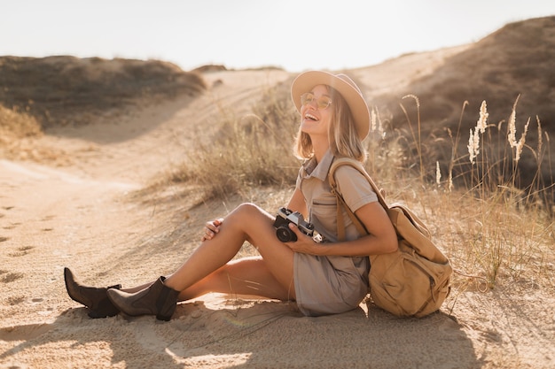 Attractive stylish young woman in khaki dress in desert, traveling in Africa on safari, wearing hat and backpack, taking photo on vintage camera