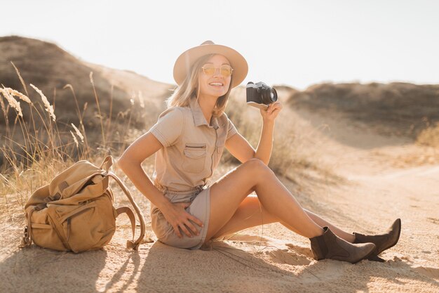 Attractive stylish young woman in khaki dress in desert, traveling in Africa on safari, wearing hat and backpack, taking photo on vintage camera