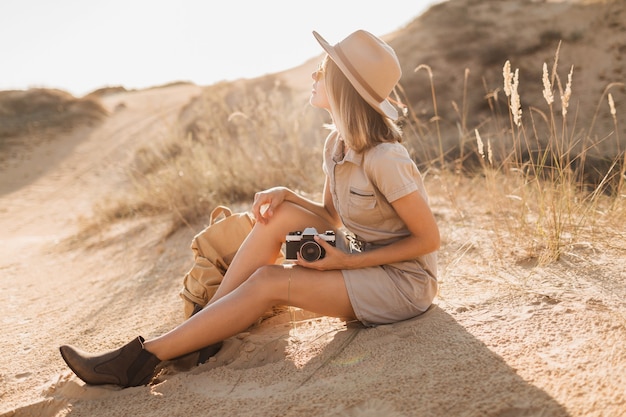 Attractive stylish young woman in khaki dress in desert, traveling in Africa on safari, wearing hat and backpack