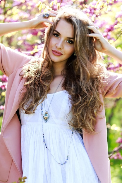 Attractive stylish young woman in light white dress, pink coat, with long hair walking in garden with blooming sakura