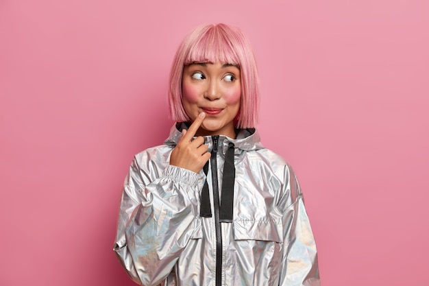 Free photo attractive teenage girl with trendy pink hairstyle, keeps index finger near lips, looks curiosly aside dressed in fashionable silver coat