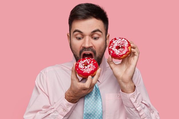 Free photo attractive unshaven businessman being hungry after hard work, bites delicious red doughnut, enjoys nice taste, wears formalwear