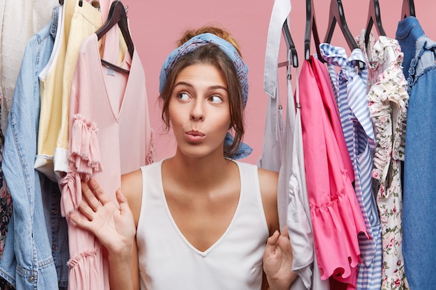 Free photo attractive woman dressed casually, looking with doubts aside while standing near hangers with clothes, thinking what to dress on business meeting with companions. woman of fashion having many clothes