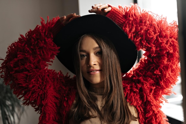 Attractive woman in fluffy jacket and widebrimmed hat is smiling sweetly Lady with straight hair posing by window