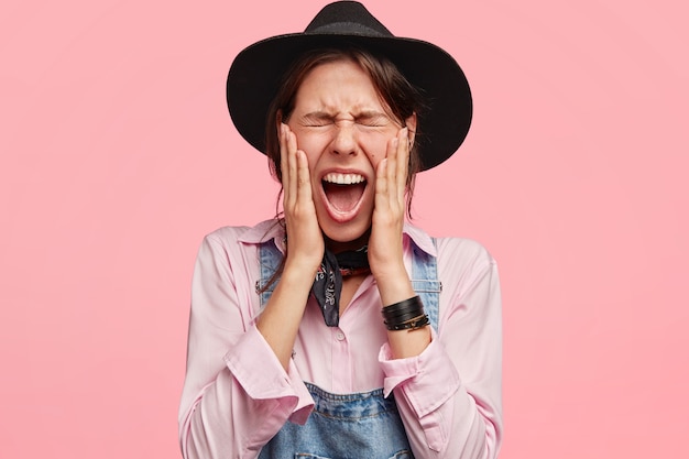 Attractive woman gardener keeps eyes shut, opens mouth widely, touches cheeks, wears hat and overalls