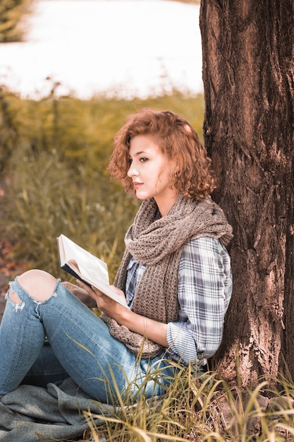 Free photo attractive woman leaning on tree and holding book in park