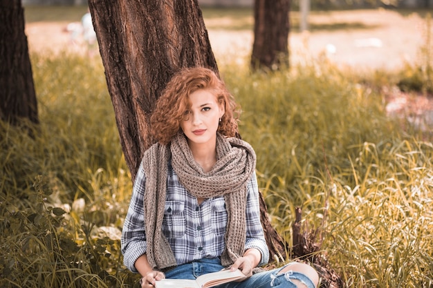 Free photo attractive woman leaning on tree and holding book in public garden