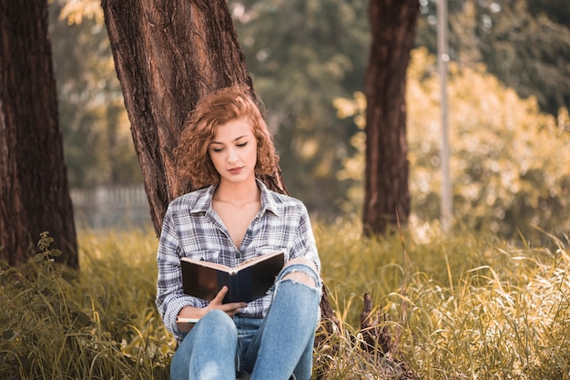 Attractive woman leaning on tree and reading book in public garden