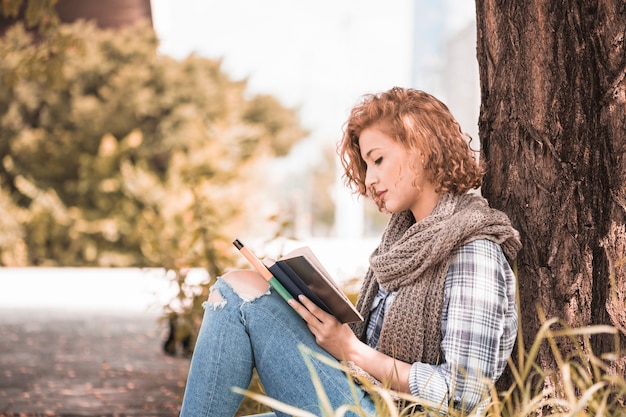 Free photo attractive woman leaning on tree and reading book in sunny park