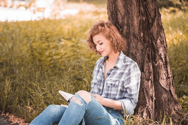 Free photo attractive woman leaning on tree and reciting book in public garden