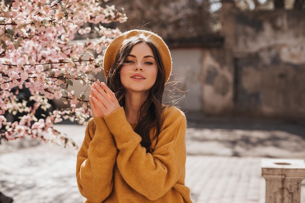 Attractive woman in orange sweater and beret poses beside sakura. Dark-haired curly lady in hat walking in sunny spring city