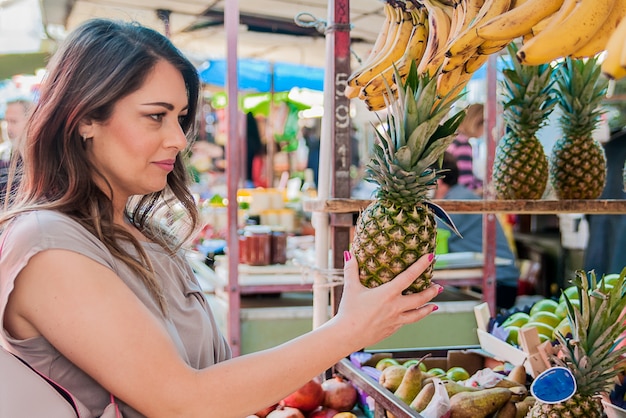 Free Photo attractive woman shopping in green market. closeup portrait beautiful young woman picking up, choosing fruits, pineapples. positive face expression emotion feeling healthy life style