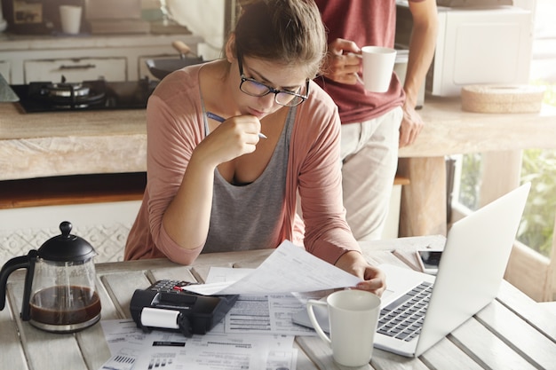 Free photo attractive woman in spectacles having serious and concentrated look holding pen while filling in papers, calculating bills, cutting family expenses, trying to save money to make big purchase