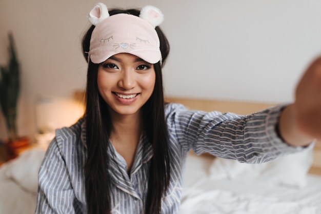 Attractive woman in striped shirt and sleep mask is smiling and taking selfie