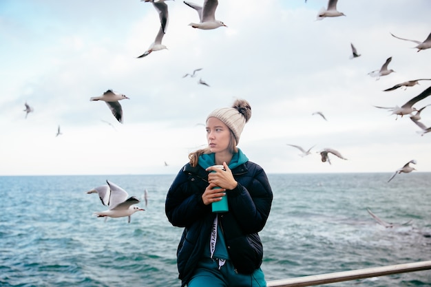 Attractive woman thinking about something, holds thermos near the ocean with seagulls