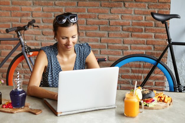 Attractive woman wearing sunglasses on her head shopping online, using high-speed internet connection, sitting in front of open laptop computer during lunch