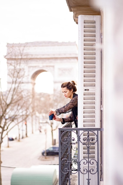 Free Photo attractive  yang woman in pajama is drinking coffee on balcony in the morning in city paris. view of the triumphal arch.