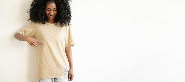 Free photo attractive young african female model wearing casual t-shirt, smiling and showing her blank t-shirt