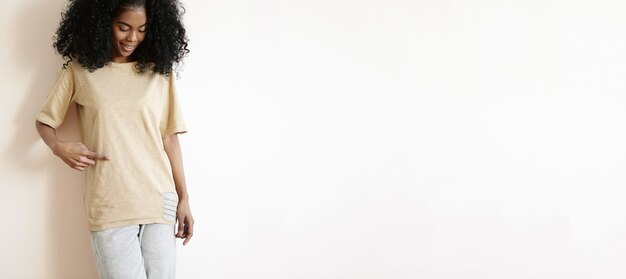 attractive young African female model with stylish curly haircut dressed casually smiling cheerfully, showing her t-shirt with finger.