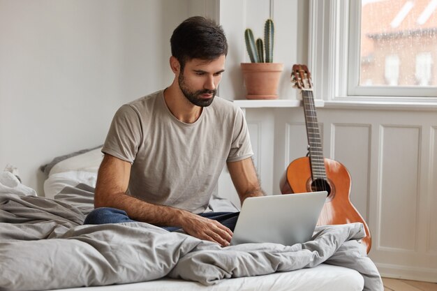 Attractive young bearded freelancer sits in front of opened laptop computer,  keeps legs folded in comfortable bed