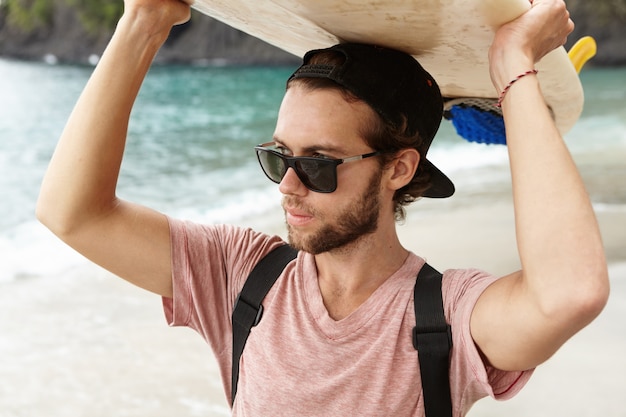 Attractive young bearded surfer in shades standing on beach at ocean