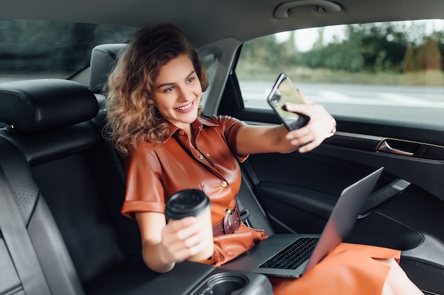 Attractive young businesswoman working on the car with a cup of coffee and holding phone while going to the work