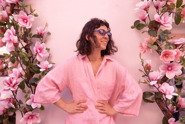 Attractive young caucasian brunette girl in pink clothes holds hands on waist looking to side on background with flowers