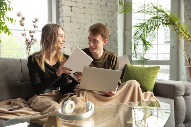 Attractive young couple using devices together, tablet, laptop, smartphone, headphones wireless. Communication, gadgets concept. Technologies connecting people in self-insulation. Lifestyle at home.