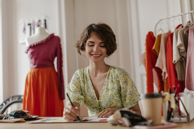 Attractive young curly woman in stylish v-neck floral dress smiles sincerely, holds pen and poses in office of fashion designer