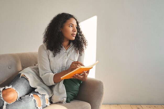 Attractive young dark skinned mulatto female with Afro hairstyle relaxing on couch at home, having pensive thoughtful look, writing down ideas for her own startup project, using pen and copybook