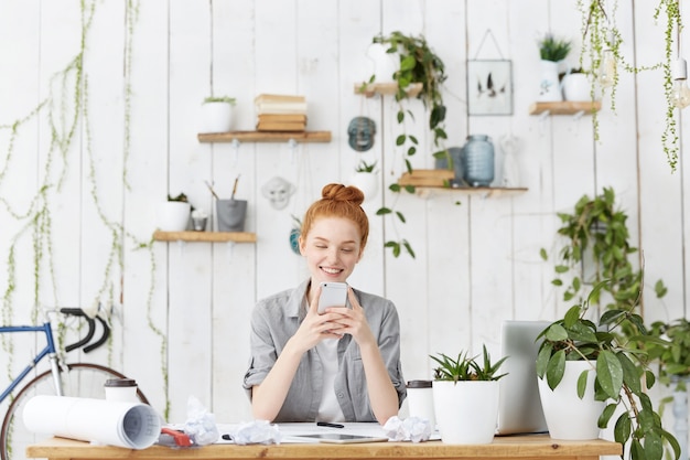 Attractive young European female architect wearing her ginger hair in bun sitting at her workspace