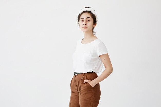 Free Photo attractive young female model with dark and wavy hair in bun, wearing white t-shirt and trousers, keeping her hands in pockets, posing against white wall with copy space for advertisment