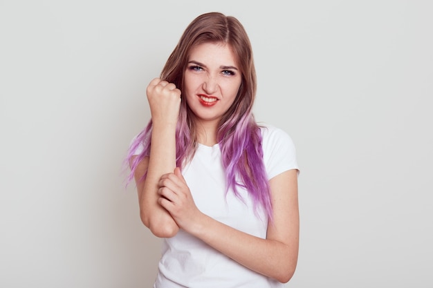 Free photo attractive young girl with lilac hair hurts her arm, touching pain hand and looking directly at camera, has serious injury, isolated over grey background.