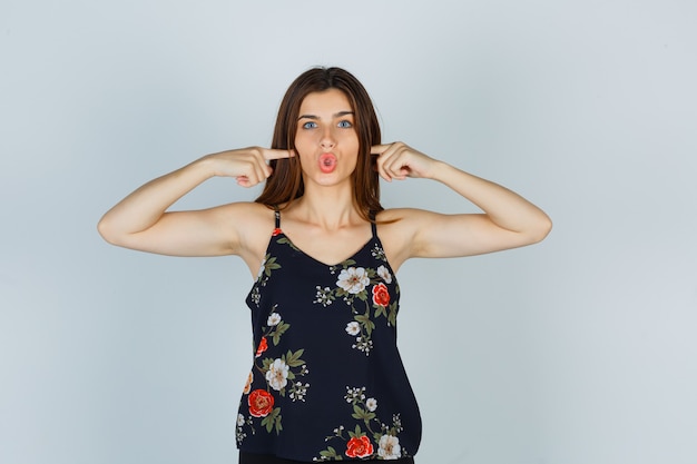 Attractive young lady pressing fingers on cheeks, keeping lips folded in blouse and looking surprised. front view.