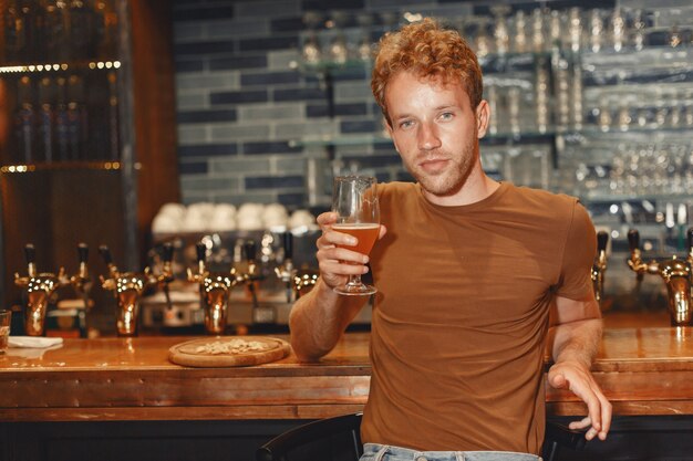 Attractive young man standing behind the bar. Man in a brown T-shirt holds a glass in his hands.