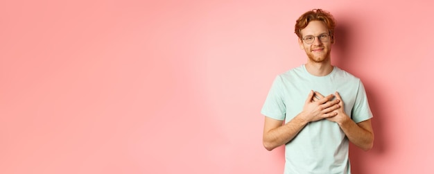 Free Photo attractive young man with ginger hair holding hands on heart and smiling grateful saying thank you e