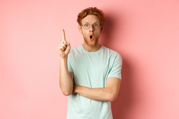 Attractive young man with red hair, raising finger in eureka gesture and pitching at idea, standing over pink background