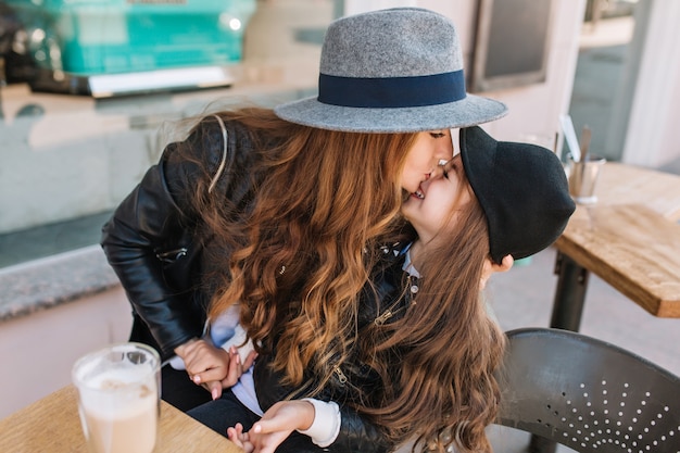 Free Photo attractive young mom wearing hat with black ribbon funny kissing her daughter in nose, while she laughing.