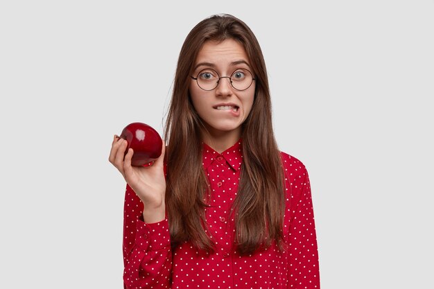 Attractive young woman bites lips lower lips, holds fresh red apple, has healthy nutrition, wears red polka dot shirt, stands against white background