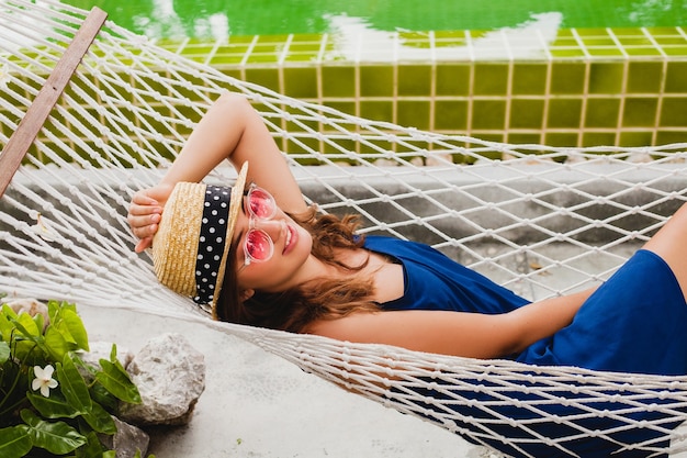 Attractive young woman in blue dress and straw hat wearing pink sunglasses relaxing on vacation lying in hammock in summer style outfit, smiling happy