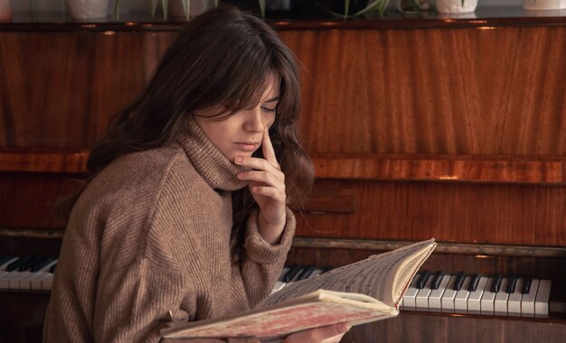 An attractive young woman in a cozy sweater sits near the piano and looks at the notes.