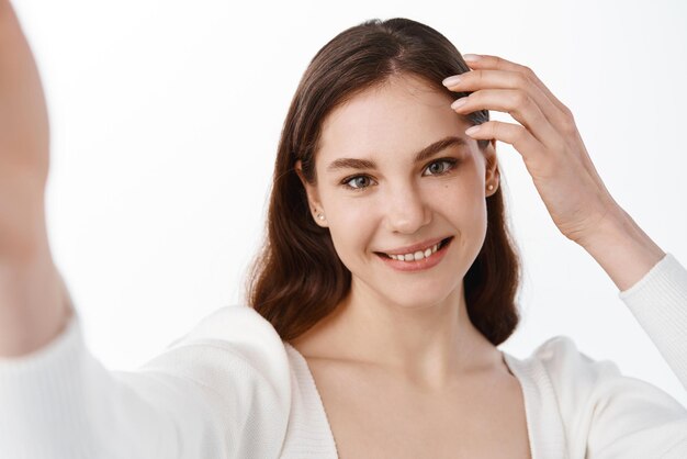 Attractive young woman holding camera in stretched hand and taking selfie making her photo on mobile phone smiling standing over white background