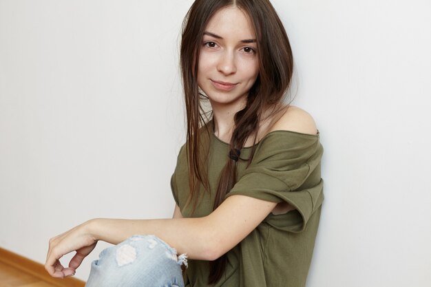 attractive young woman model wearing stylish clothes, sitting on floor, leaning back on white wall, having friendly and relaxed expression on her pretty face