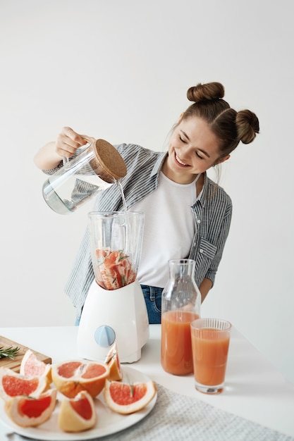 Free photo attractive young woman smiling adding water in blender with grapefruit pieces and rosemary. healthy diet food nutrition.