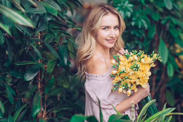 An attractive young woman standing near plants holding delicate yellow freesia in hand