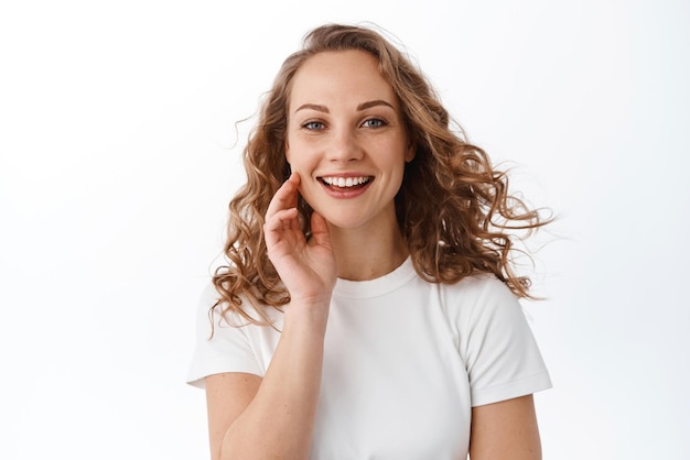 Attractive young woman touching natural face without make up smiling pleased and looking at camera standing in tshirt against white background