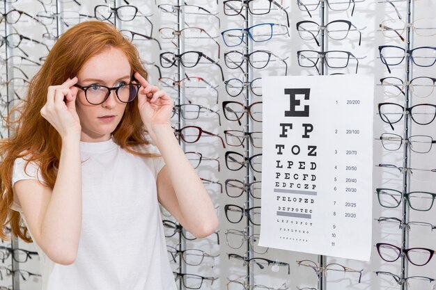 Attractive young woman wearing eyeglasses standing neat snellen chart in optica