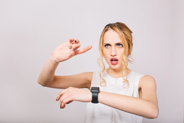 Free Photo attractive young woman with curly hair wearing stylish blouse looks at black wristwatch and plans day. charming blonde lady in white tank top remembers list of cases for today and calculates time.