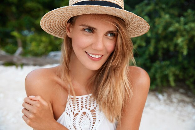 Attractive young woman with dreamy positive expression, dressed in fashionable hat and summer white dress, looks thoughtfully into distance, plans something for next day, has good rest abroad